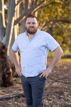 bearded man with hands on hips standing outdoors with trees in background