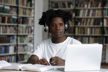 Serious African student guy sit at table look at laptop makes assignment, listen audio through...