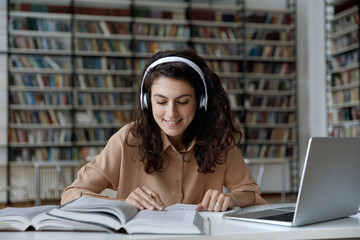 Young 20s attractive girl in headphones sit at table with textbooks and laptop, read topics, prepare essay or summary. Diligent student learns, gain new knowledge in college library. Education concept
