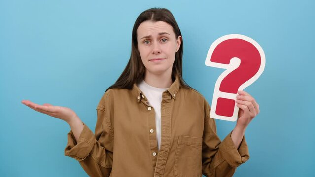 Portrait of pensive young caucasian woman 20s old year holding red paper question mark, thinks about tasks, looking away, wearing brown shirt, posing isolated over blue color background wall in studio