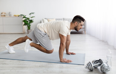 Handsome young Arab guy in sportswear exercising on yoga mat, doing domestic fitness, working out...