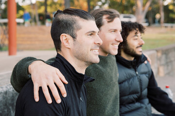 Close up shot of 3 smiling interracial men sitting on a field, hands on shoulders