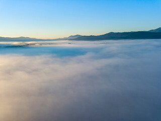 Fototapeta premium Flight over fog in Ukrainian Carpathians in summer. Mountains on the horizon. A thick layer of fog covers the mountains with a continuous carpet. Aerial drone view.