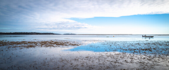 Soft seascape with white clouds and blue sky reflected on the grassy bay with a mooring boat.