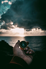 Pov of man hand holding navigational travel compass to find direction and next destination place. Concept of travel people and exploring. Beach and ocean with sdramatic ky light in background © simona