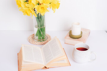A cup tea, book, candle and daffodils in glass vase. Cozy home, reading or studying concept
