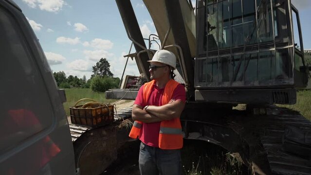 Outdoor portrait of chief in sunglasses retroreflective vest and construction helmet stands near undercarriage of caterpillar excavator