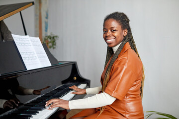 Portrait of African young woman smiling at camera while sitting at piano, she happy to learn...