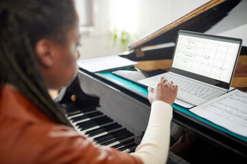 Rear view of African woman looking at sheet music on monitor of her laptop while learning to play...