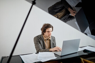 Serious student concentrating on his work on laptop while sitting at grand piano, he having online...