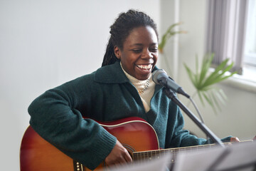 African young woman singing a happy song to microphone and playing the guitar while sitting in...