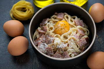Middle close-up of pasta carbonara served with an egg yolk in a black bowl, horizontal shot on a dark-brown stone surface