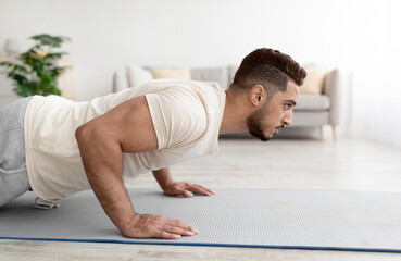 Side view of young athletic Arab man doing push-ups on sports mat at home, copy space - obrazy, fototapety, plakaty