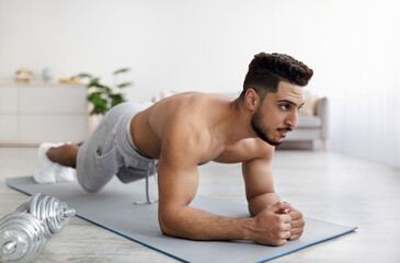 Shirtless young Arab man standing in elbow plank on yoga mat, exercising abs muscles on domestic training indoors
