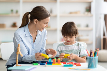 Cute little boy playing logical game with positive woman teacher, studying together at educational playroom, free space