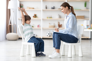 Cute talkative little boy telling story to professional woman psychologist during consultation, therapist taking notes