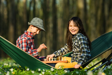 Two kids boy and girl playing chess while sitting in a folding hammock in a forest or park. Spring...