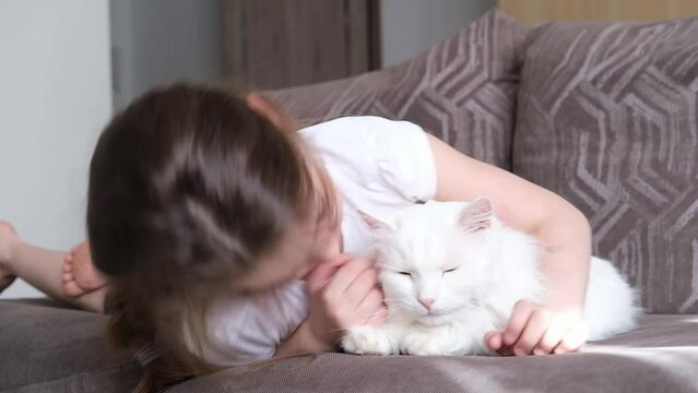 A little happy white girl lies on the sofa in the living room, hugs a white fluffy cat and smiles, closing her eyes.