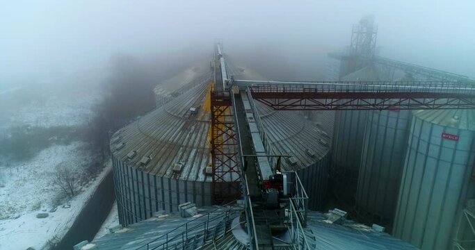 Flying over the tops of metal granary elevators. Huge tanks standing close and connected by supports. Foggy winter day backdrop.