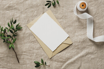 Blank white paper card with envelope, green branches, silk ribbon on linen cloth background. Wedding invitation template.