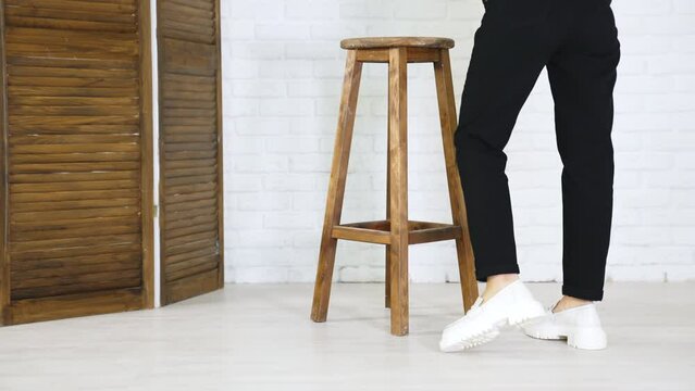 Model in black jeans demonstrating white massive shoes. Lady turns around to show the fashionable footwear. Wooden tabouret at backdrop.