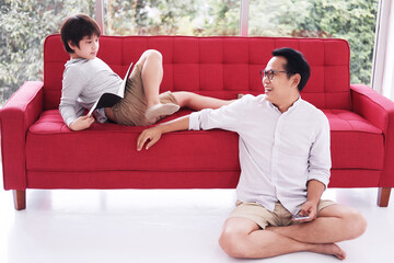 Smiling Asian father is enjoying with his son. Boy is lying and reading a book on red sofa in...