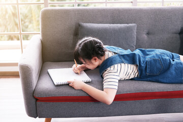 A little girl is lie down and enjoying drawing on the sofa. She is working for homework on holiday at her house. Education and homeschool Concept