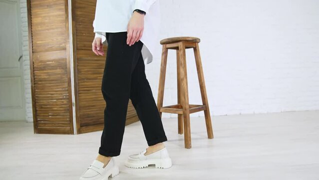 Lady in white shirt and black jeans demonstrates white fashionable shoes. Woman walks in plain loafers with massive soles back and forth near wooden tabouret.