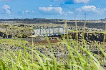 Metal bridge over the American and Eurasian tectonic plates in sunshine. Volcano landscape on the Reykjanes Peninsula of Iceland. View through green blades of grass in the foreground