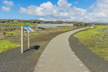 Pathway with a sign pointing to a metal bridge on the Reykjanes Peninsula of Iceland. Lava rocks...