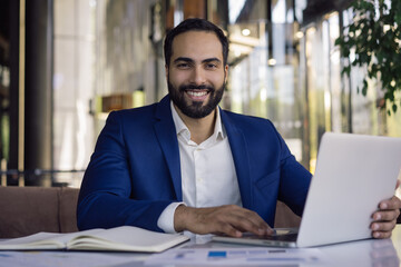 Handsome smiling Moroccan businessman using laptop computer working online sitting in modern...