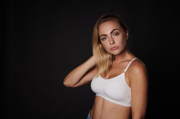 Fototapeta na wymiar Beautiful slim body Caucasian woman, fit and muscular build young fitness girl in white top confidently looking at camera, isolated over black background with copy ad space