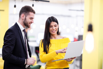 Beautiful Caucasian dark haired girl in yellow posing with businessman wearing suit. Happy colleagues with a laptop in a modern office.