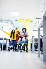 Colleagues having fun on work at the office. Girls pushing boys on chairs with wheels. Multiethnic group of successful start up company.