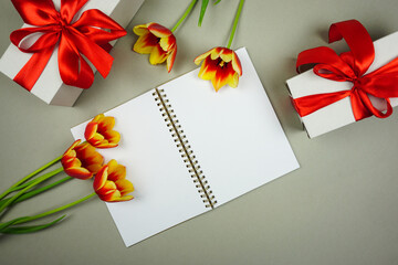 On a gray background there is an open notebook for writing and there are red and yellow tulips and boxes with gifts
