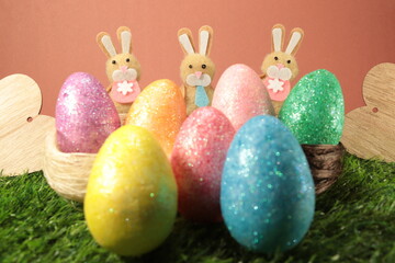Three easter bunnies spaced evenly apart behind colorful easter eggs against an orange-reddish background with wooden hearts placed on either side.