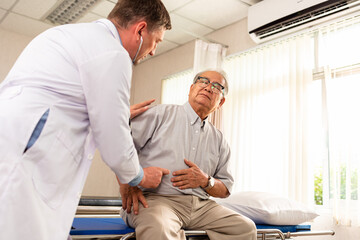 Asian senior male patient sitting on examination bed pointing at the stomach that pain to a Caucasian male doctor.Elder man showing stomach ache pain to doctor in clinic. - obrazy, fototapety, plakaty