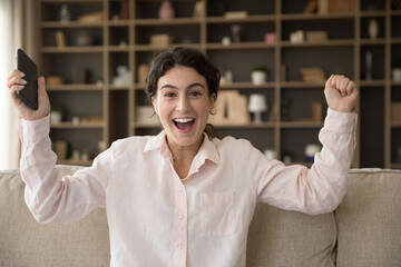 Obraz premium Excited surprised mobile phone user girl receiving happy good news, making winner triumph gesture, raising hands, shouting yes, holding smartphone, laughing, smiling, shouting at camera