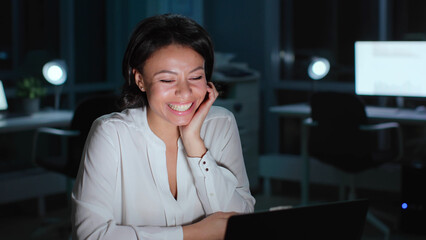 Happy African-American businesswoman look at laptop screen and smile in dark office