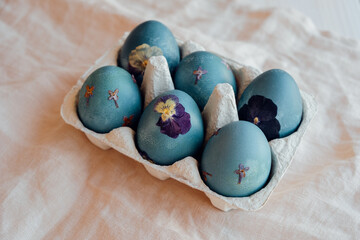 Naturally dyed cyan blue pressed flower Easter eggs in egg box on neutral beige tablecloth background. Handmade craft natural decoration.