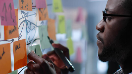 Close up of pensive African-American businessman look at glass board with sticky notes
