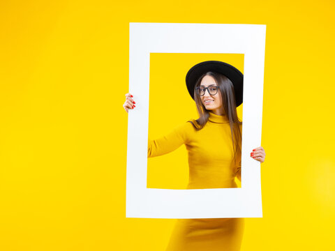 Beautiful woman holds a frame and smiles - isolated over yellow. ?heerful girl in the picture. Woman looks into the camera and smiles. Girl poses with a white frame. Lady in a hat and a yellow dress.