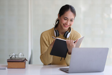 An Asian woman writes a memo of learning from the Internet. Happy woman working at home holding a notebook writing notes. Work from home concept.
