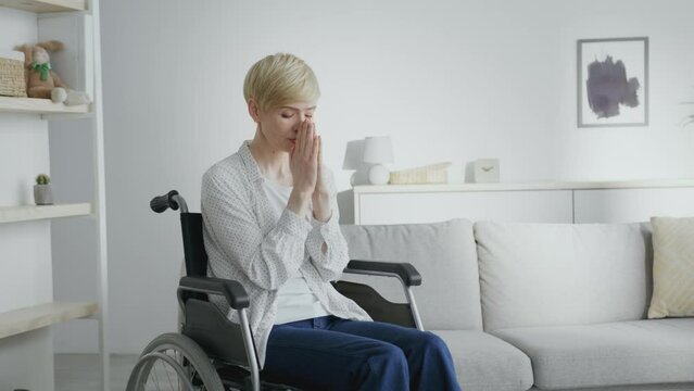 Mature hopeless woman in with spinal injury praying to Gao, sitting in wheelchair at home interior, empty space