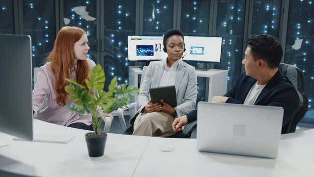 Business meeting in data center. Multi-ethnic experienced staff of IT workers communicating about business plans. Cyber security office. Server racks. Teamwork.