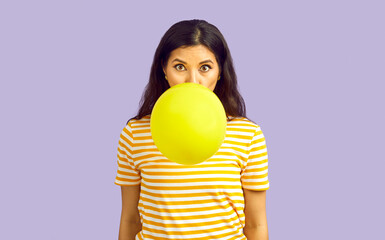 Woman blowing up balloon. Pretty long haired brunette girl with increased lung capacity standing isolated on lilac background, inflating yellow balloon and looking at camera with funny face expression