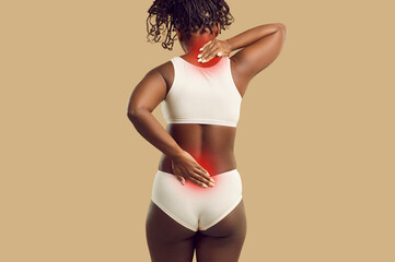 Plus size black woman in underwear who suffers from discomfort and backache and needs spine physiotherapy touching painful inflamed neck and lower back area, backside view isolated on color background