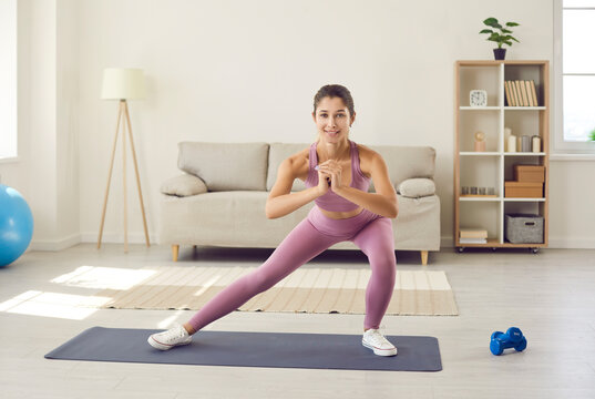 Happy beautiful slim woman enjoying routine fitness workout at home. Active young Caucasian lady in modern sports leggings standing on gym mat in living room, smiling and doing side lunge exercise