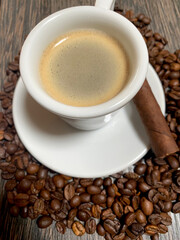 Closeup of espresso cup with foam and a cigar on plate and coffee roasted beans on wooden table
