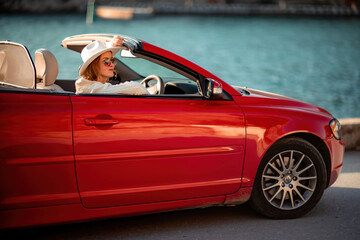 Outdoor summer portrait of stylish blonde woman driving red car convertible. Fashionable attractive woman with blond hair in a white hat in a red car. Sunny bright colors taken outdoors